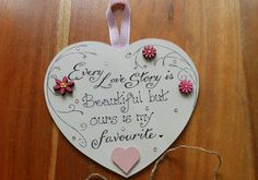 a heart shaped sign hanging on a wooden table with some twine and pink flowers