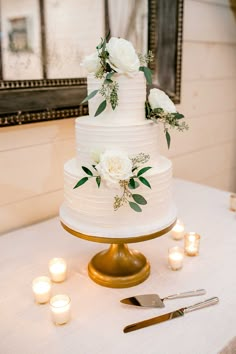 a wedding cake with white flowers and greenery sits on a table next to candles