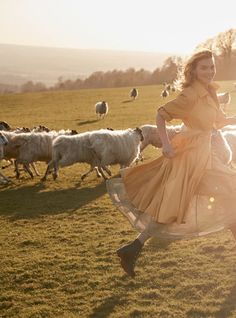 a woman in a yellow dress is running with some sheep on the grass and sun shining down