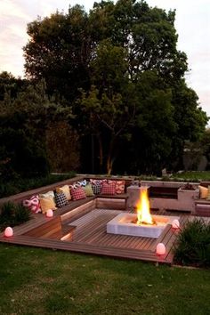 an outdoor fire pit surrounded by wooden decking and pillows on the ground with trees in the background