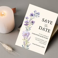 a wedding card next to a lit candle and some dried flowers on a gray surface