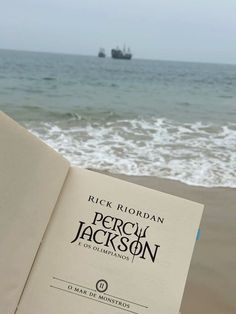 an open book sitting on top of a beach next to the ocean with a ship in the background