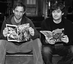 two men sitting on a couch holding comic books in their hands and looking at the camera