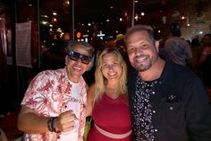 two men and a woman posing for a photo at a bar with people in the background