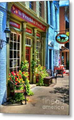 an old fashioned store front with flowers in the window and bench on the sidewalk metal print