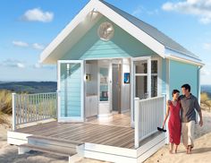 a man and woman walking towards a tiny blue house on the beach with stairs leading up to it