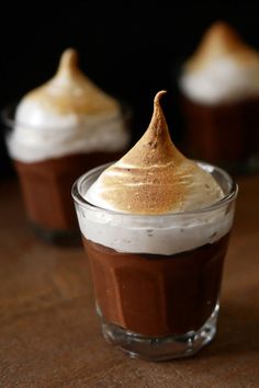 three desserts in small glass cups with whipped cream and chocolate on top, sitting on a wooden table