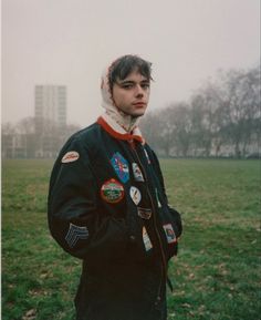 a man standing in the middle of a field with patches on his jacket and scarf around his neck