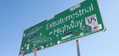 a green and white street sign with stickers on it's side in front of a blue sky