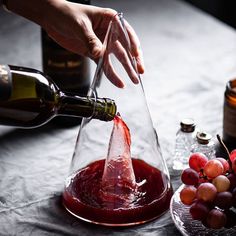 a person pouring red wine into a glass with grapes on the table next to it