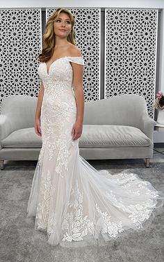 a woman standing in front of a couch wearing a wedding dress with an off the shoulder neckline