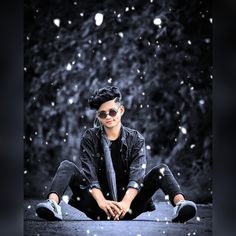 a young man sitting on the ground in front of trees and snow flakes, wearing sunglasses