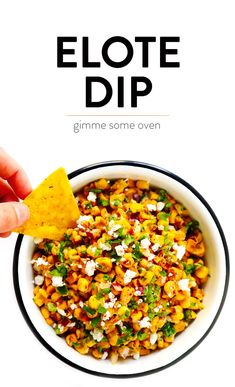 a hand holding a tortilla chip over a bowl filled with mexican corn and feta cheese