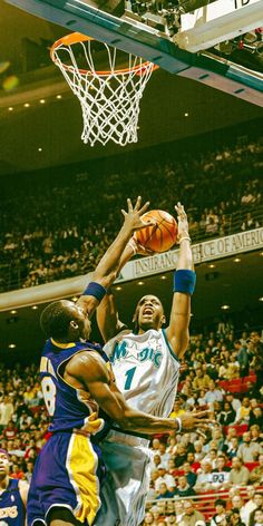 two men playing basketball in front of an audience at a sporting event, one is jumping up to grab the ball