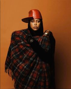 a woman wearing a red hat and plaid ponchy wrapped in a blanket with her hands on her hips