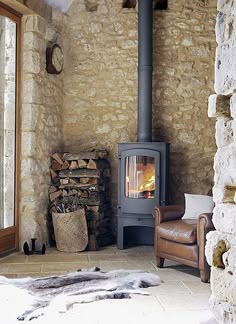 a wood burning stove in a stone walled room with a leather chair and firewood stacked on the floor