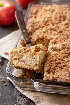 an apple crumb cake in a glass dish with one slice cut out and two apples behind it