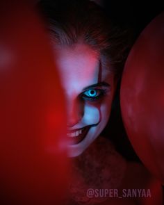 a woman with blue eyes and clown makeup looks at the camera from behind red balloons