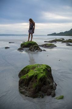 a woman standing on some rocks covered in green mossy algae at the ocean's edge