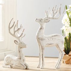 two white deer figurines sitting next to each other on a wooden table with flowers in the background