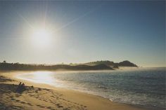 two people sitting on the beach watching the sun rise over an island in the distance