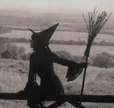 an old photo of a woman dressed as a witch holding a broom and sitting on a fence