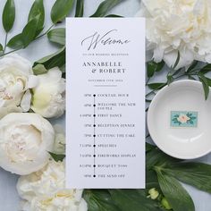 a wedding program with white flowers and green leaves on the table next to a plate