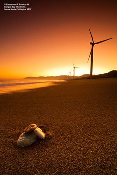 the sun is setting behind windmills on the beach with rocks in front of them