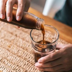 a person pouring something into a glass on top of a wooden table next to a brown mat