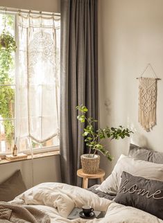 an unmade bed in front of a window with plants on the side table next to it