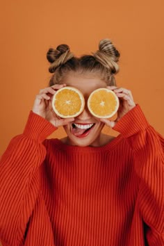 a woman holding two halves of oranges up to her eyes with both hands and smiling