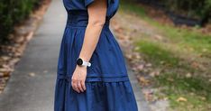 a woman in a blue dress is standing on the sidewalk with her hands behind her back