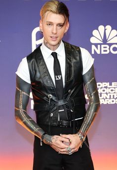 a man with tattoos on his arm wearing a black leather vest and white shirt is standing in front of a purple wall