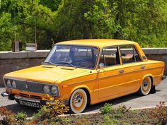 an old yellow car parked in a parking lot next to some bushes and trees on the side of the road