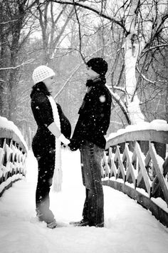 two people are standing on a bridge in the snow, holding hands and looking at each other