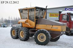 an orange and brown truck parked in front of a building on snow covered ground with other vehicles behind it