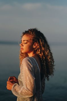 a woman with long curly hair standing by the water looking off into the distance at sunset