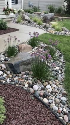 a garden with rocks and flowers in the front yard, next to a house on a hill