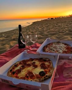 two pizzas in boxes sitting on the beach with wine glasses and bottle next to them