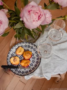 a plate with peaches on it next to some wine glasses and pink peonies