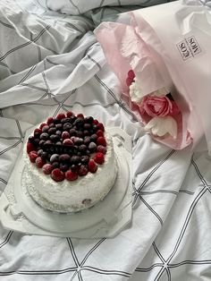 a white cake with berries on it sitting on a bed next to a pink rose