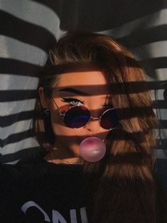 a woman with sunglasses blowing bubble in front of her face and shadows on the wall behind her