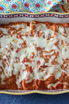 a casserole dish filled with meat and cheese on a blue tablecloth next to a colorful cloth