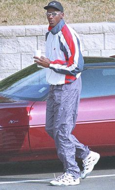 a man standing in front of a red car holding a drink and looking at his cell phone