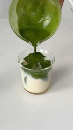 a person pouring green liquid into a small glass bowl on top of a white table