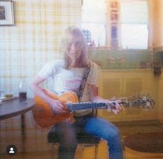 a woman sitting on a chair playing an orange guitar in front of a window with yellow curtains