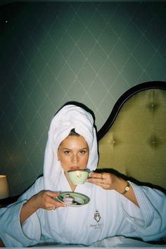 a woman sitting in bed drinking from a cup and saucer while wearing a white robe