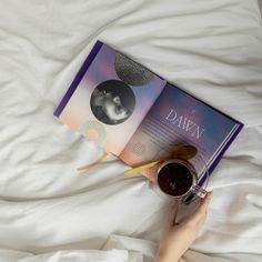 a person laying in bed reading a book and holding a cup of coffee next to it