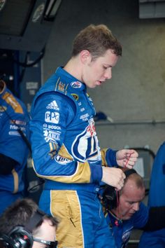 a man in a blue and yellow racing suit standing next to another man wearing headphones