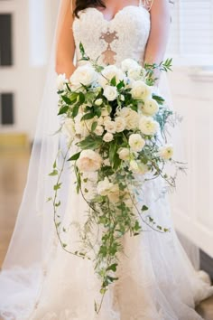 a woman in a wedding dress holding a bouquet of white flowers and greenery on the floor